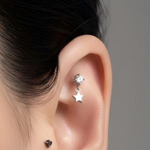May include: A silver earring with a dangling star charm and a small, clear gemstone. The earring is worn on the upper ear. A small, black stud earring is also visible on the earlobe.
