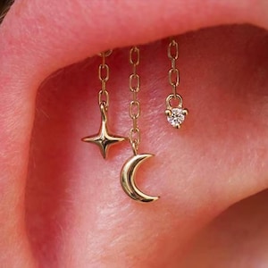 Pode incluir: Close-up de um piercing de orelha dourado com três pingentes pendurados: uma estrela de quatro pontas, uma lua crescente e um pequeno pingente com uma única pedra preciosa transparente. Os pingentes são suspensos por correntes delicadas.