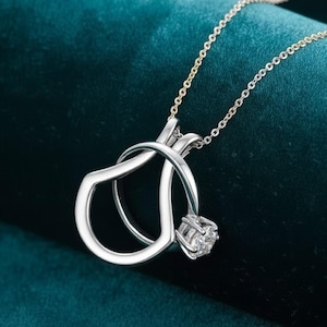 Puede incluir: Un collar con colgante plateado con una gema con forma de diamante engastada en un diseño de anillo estilizado. El colgante cuelga de una delicada cadena, creando un aspecto sofisticado. El collar se muestra sobre un fondo verde azulado oscuro.