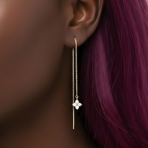 May include: A close-up of a gold earring. The earring has a fine chain that hangs from the ear. At the base of the chain is a four-petal flower design with small, clear gemstones. A small gold bar hangs below the flower.