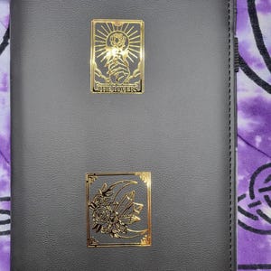 May include: Black leather journal with two gold-colored metal embellishments. The top embellishment features a sun and hands with the text "THE LOVERS". The bottom embellishment has roses, a crescent moon, and crystals. The journal has a stitched edge.
