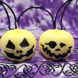 May include: Two round, yellow candles carved with jack-o'-lantern faces. The black facial features include triangular eyes and a jagged mouth. The candles sit on a purple and white patterned fabric.