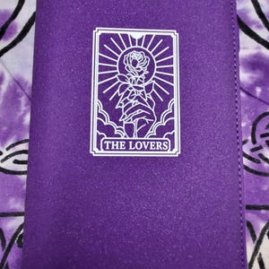 May include: A rectangular purple wallet with a textured surface. Centered on the front is a white graphic print of a hand holding a rose beneath a sunburst, enclosed in a rectangular frame with the text THE LOVERS printed at the bottom.