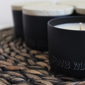 May include: Black candle in a black jar with a wooden lid. The candle has a white wax top and the jar has the text "PWB MANdle" printed on the front.