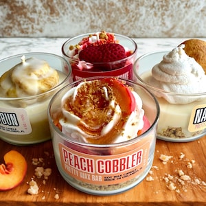 May include: Four dessert-themed soy wax candles in clear glass jars. The candles are decorated to resemble a cinnamon bun, strawberry cheesecake, peach cobbler, and a banana pudding. The peach cobbler candle has a label that reads "Peach Cobbler Pretty Wax Bar".