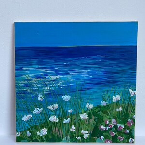 Puede incluir: Una pintura de un paisaje marítimo con flores blancas en primer plano y un océano azul en el fondo. El cielo es de un azul brillante.