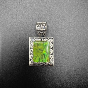May include: A rectangular pendant with a vibrant green and yellow gemstone set in a silver-toned frame. The frame features an ornate, scrolling design around the stone, and a decorative bail at the top for a chain.