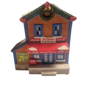 May include: A miniature Christmas village building with a brown wooden facade, blue trim, and a red awning. The building is a True Value Hardware store with Coca-Cola branding. A green wreath and gold bells adorn the roof.