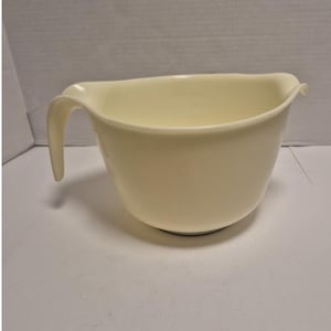 May include: Cream-colored mixing bowl with a handle. The bowl has a rounded shape and a pouring spout. The handle is curved and attached to the side of the bowl. The bowl is made of a smooth, solid material.