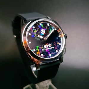 May include: A black wristwatch with a silver case and a black rubber strap. The watch face has a black background with a colourful pixelated design and the text "STC Quanta".