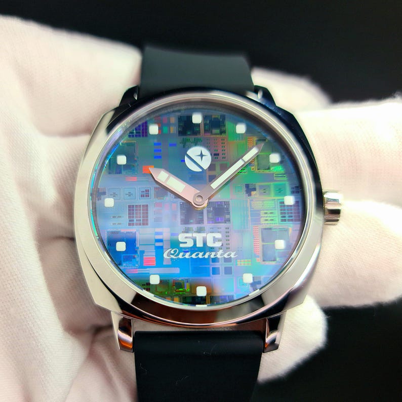 May include: A silver wristwatch with a black rubber band. The watch face has a circuit board design with a white background and a silver star in the center. The text "STC Quanta" is printed on the watch face.