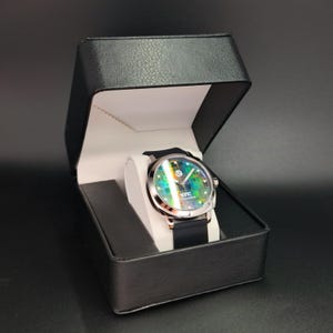 May include: A black watch with a green, blue, and yellow patterned face. The watch is in a black box with a white interior.