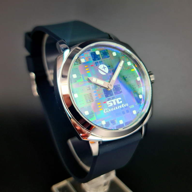 May include: A silver watch with a blue silicone band. The watch face has a circuit board design with a white star and the text "STC Quanta" in black.