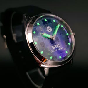 May include: A silver watch with a black band and a green glow-in-the-dark face. The face has a circuit board design and the text "STC Quanta".