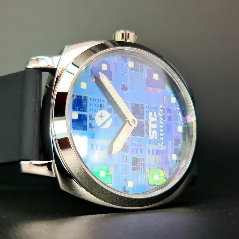 May include: A silver wristwatch with a blue face and a black rubber band. The face of the watch has a circuit board design and the text "STC Quanta" printed on it.
