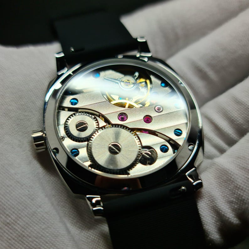 May include: A silver watch with a black leather band. The watch face is clear and shows the inner workings of the watch.