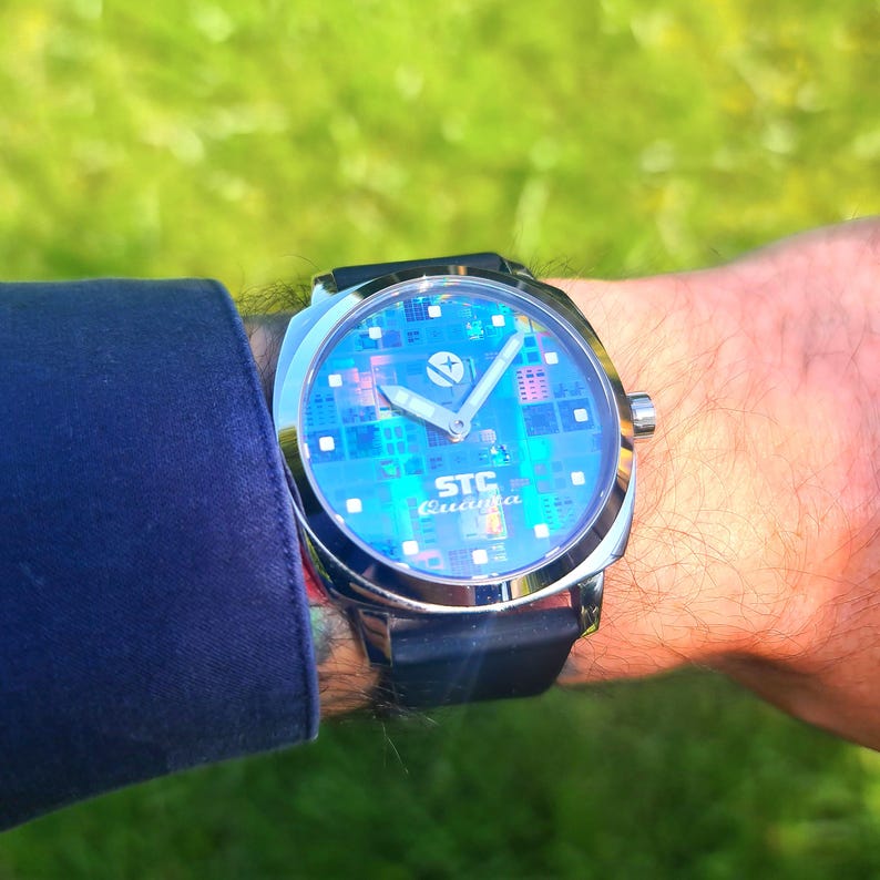 May include: A silver watch with a black band and a blue face. The face has a circuit board design and the text "STC Quanta" printed on it.