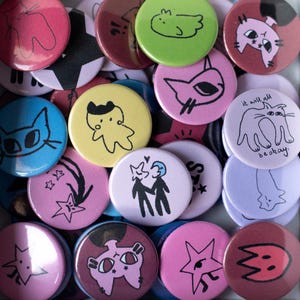 May include: A collection of circular pin-back buttons in a square box, featuring various colourful designs including cats, stars, and figures. One button displays a cat illustration with the text 'it will all be okay' written in black ink.