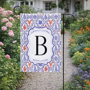 May include: A garden flag with a white background featuring a blue and red nautical pattern. The flag has a white central panel with a large black letter "B" inside a decorative blue frame. The flag is displayed on a black metal stand.
