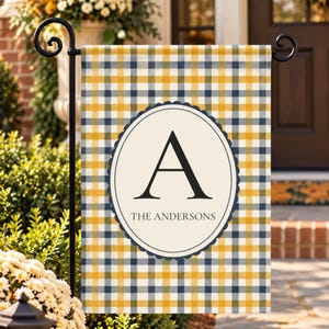 May include: A rectangular garden flag featuring a yellow, navy blue, and white plaid pattern. In the centre, a cream-coloured oval with a scalloped navy border displays a large black letter A above the text THE ANDERSONS in black font.