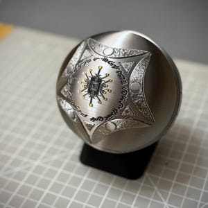 May include: A spherical metal object with a brushed silver finish, featuring a central engraved star design. The star contains a black and gold circuit board pattern and is surrounded by circular indentations and etched text.