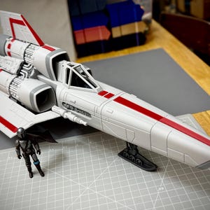 Puede incluir: Una maqueta de nave espacial blanca y roja con un diseño detallado, con motores y alas. Una pequeña figura de acción está cerca. La nave espacial tiene un cuerpo largo y aerodinámico con rayas y detalles rojos.