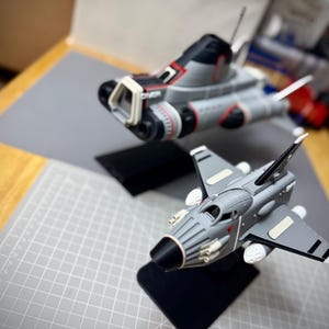 Könnte beinhalten: Zwei graue und schwarze Modellraumschiffe mit roten und weißen Akzenten. Eines ist ein größeres Shuttle-Design, das andere ein kleinerer Jäger. Beide stehen auf schwarzen Ständern, vor einer grauen Schneidematte und einer Holzoberfläche.