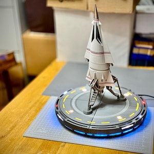 May include: A white and silver model rocket ship with red and blue horizontal stripes stands on a circular gray launch pad. The base features illuminated yellow and blue lights around its perimeter, with a metallic pointed nose cone at the top.