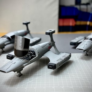 May include: Three gray model airplanes with black accents are arranged on a grid surface. The planes have a sleek design with wings, a cockpit, and a tail fin. The models are likely for display or hobby purposes.