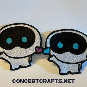 以下が含まれることがあります： 黒い目と青い瞳を持つ白い刺繍のロボットのパッチが2つ。それぞれのロボットには、ピンクとブルーのハートが側面についています。