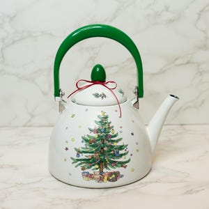 May include: White Christmas-themed teapot with a green handle and lid. The teapot features a decorated Christmas tree design with presents and ornaments. The handle is a curved, bright green color.