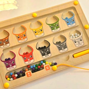 A wooden toy with a maze for sorting colorful marbles. The maze has eight sections, each with a different color: brown, orange, yellow, green, blue, white, gray, and black. Each section has a number and a cartoon image of a bucket. The maze also has a section for counting marbles with a plus and equals sign. Interactive Montessori math toy for toddlers, color sorting and counting board to build early STEM skills