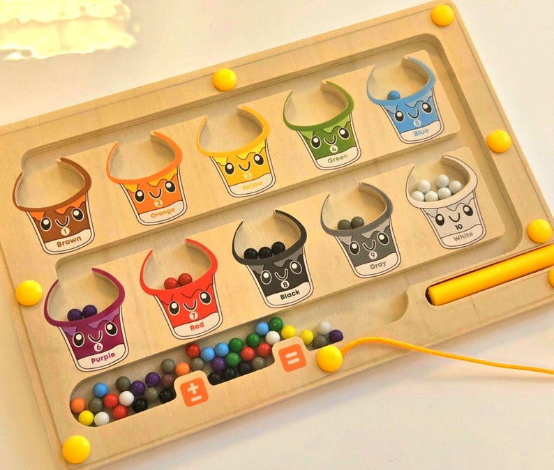 A wooden toy with a maze for sorting colorful marbles. The maze has eight sections, each with a different color: brown, orange, yellow, green, blue, white, gray, and black. Each section has a number and a cartoon image of a bucket. The maze also has a section for counting marbles with a plus and equals sign. Interactive Montessori math toy for toddlers, color sorting and counting board to build early STEM skills