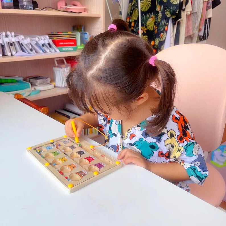 A wooden toy with colorful beads and a yellow string. The toy has a grid of 12 compartments with different colored beads in each compartment. The string is threaded through the beads and can be moved around the grid. Montessori toddler busy board packaged for gifting, educational interactive kids toy for birthday or Christmas gift