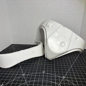 May include: Two white molded plastic armor components with smooth, curved surfaces. One piece is a wide, arched section, while the other is a larger, contoured plate featuring recessed details and raised ridges, designed for a costume assembly.