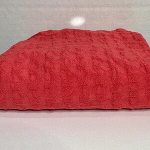 May include: A folded rectangular textile piece in a vibrant coral red hue. The fabric features a consistent, raised waffle-knit texture throughout its surface, creating a grid-like pattern of small, square indentations across the entire material.