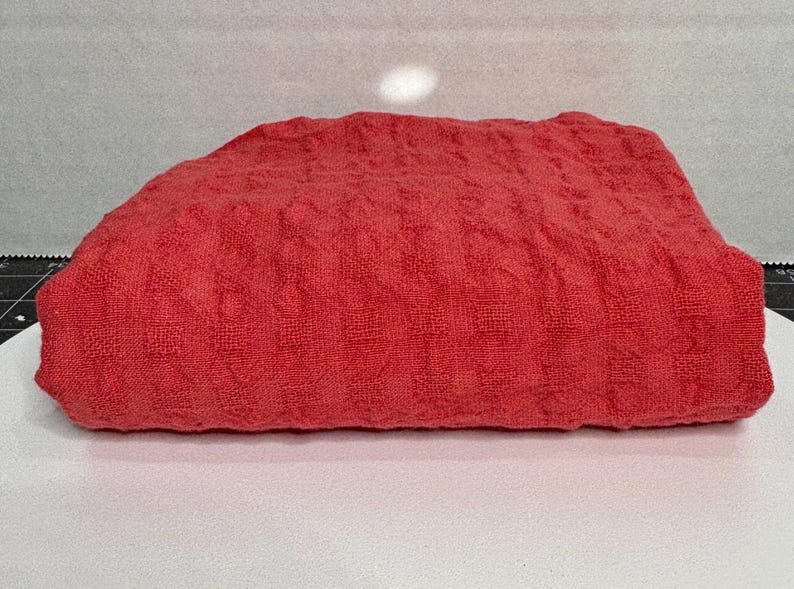 May include: A folded rectangular textile piece in a vibrant coral red hue. The fabric features a consistent, raised waffle-knit texture throughout its surface, creating a grid-like pattern of small, square indentations across the entire material.