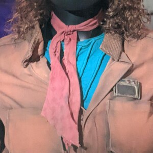 May include: A brown jacket with a textured collar and a rectangular metallic detail on the chest pocket. Underneath, a bright blue shirt is visible. A dusty rose-colored fabric neck tie with frayed edges is knotted around the neck area.