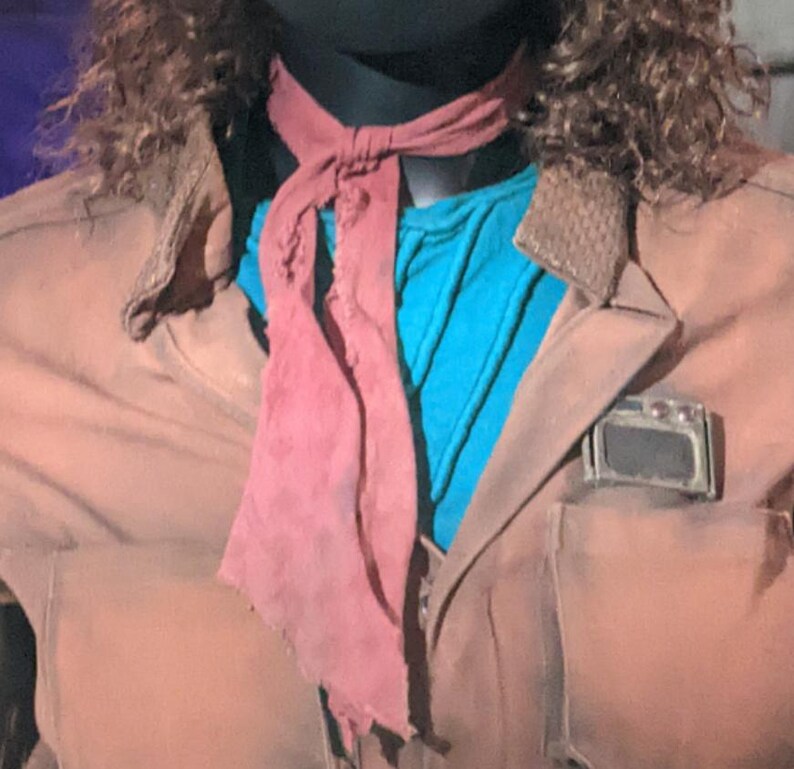 May include: A brown jacket with a textured collar and a rectangular metallic detail on the chest pocket. Underneath, a bright blue shirt is visible. A dusty rose-colored fabric neck tie with frayed edges is knotted around the neck area.
