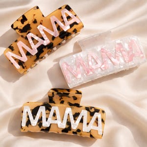Puede incluir: Tres pinzas para el pelo con la palabra "MAMA" en blanco o rosa. Una pinza tiene un estampado de carey, otra es blanca con letras rosas y la tercera tiene un estampado de carey.