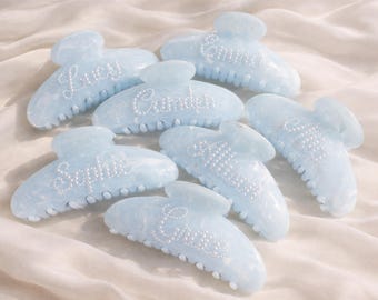 May include: Seven light blue hair claw clips with white pearl-like embellishments and names like "Lucy" and "Emma". The clips are arranged on a soft, white fabric, showcasing their marbled design.