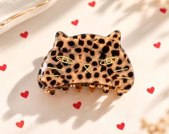 May include: A cat-shaped hair claw clip featuring a brown and black leopard print pattern. The clip has gold-toned metal line art detailing that forms two eyes, a small nose, and three whiskers on each side of the face.