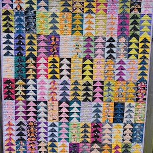May include: A rectangular quilt featuring a repeating pattern of vertical columns composed of small fabric triangles. The quilt displays a diverse array of colors including black, orange, purple, yellow, pink, and teal, with various printed motifs.
