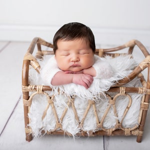 May include: A newborn baby sleeping in a wicker basket lined with a white fluffy blanket. The baby is wearing a white knitted onesie.