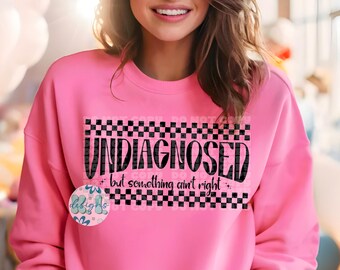 Pode incluir: Camisola rosa com acabamento em xadrez preto e o texto "Undiagnosed but something ain't right".