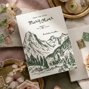Könnte beinhalten: Eine weiße Save-the-Date-Karte mit einer grünen Tuschezeichnung eines Gebirges und eines Kiefernwaldes. Der Text lautet: SAVE THE DATE, Marie & Oliver, SAT SEPT 30 20XX und Invitation to follow. Die Karte ist von Blumen und Briefmarken umgeben.
