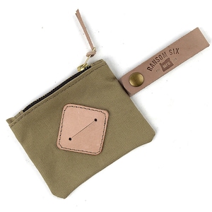 May include: A small, green canvas pouch with a zipper closure and a leather strap. The strap has a brass button and a leather tag that reads "Ransom Six Mfg. Est. 2016".