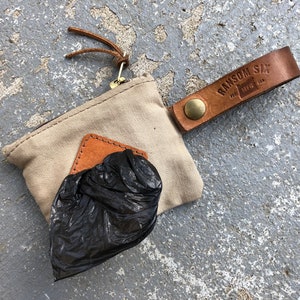 May include: A beige canvas pouch with a brown leather strap and a metal zipper. The pouch has a brown leather patch with the text "RANSOM SIX" and "MFG. USA". The pouch is filled with a black plastic bag.