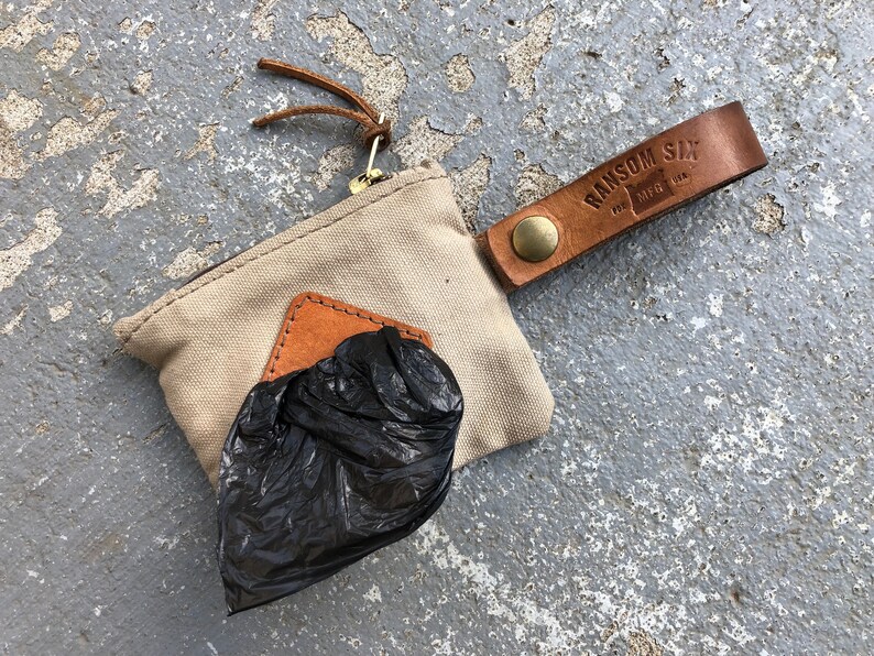 May include: A beige canvas pouch with a brown leather strap and a metal zipper. The pouch has a brown leather patch with the text "RANSOM SIX" and "MFG. USA". The pouch is filled with a black plastic bag.