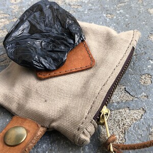 May include: A beige canvas bag with a brown leather flap and a zipper closure. A black plastic bag is on top of the flap. The bag has a brown leather strap with a brass button.